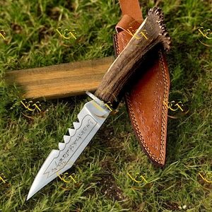 Puede incluir: Un cuchillo de caza con una hoja dentada y un mango de asta. El cuchillo está en una funda de cuero marrón con costuras decorativas. La hoja tiene texto grabado. El cuchillo descansa sobre un bloque de madera y hierba verde.