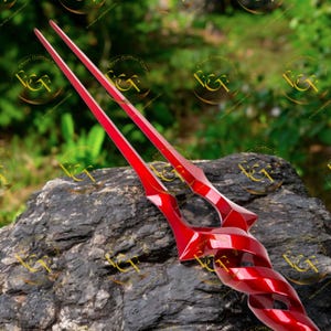 May include: Two bright red, stylized spear-like objects with a glossy finish. The design features sharp angles and a twisted central section. The objects are resting on a dark, textured rock. The background is blurred green foliage.