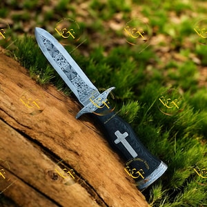 May include: A decorative dagger with an ornate, engraved blade and a black handle. The blade features detailed patterns, and the handle has a white cross design. The dagger rests on a wooden surface with a green, grassy background.