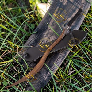 May include: A curved sword with a brown blade and a black handle wrapped in black material. The sword rests on a black sheath, placed on a weathered wooden surface, surrounded by green grass.
