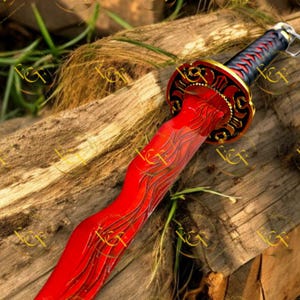 May include: A decorative red sword with a curved blade and intricate gold and black detailing on the hilt. The blade features etched designs. The handle is wrapped in black and red. The sword rests on a wooden surface.