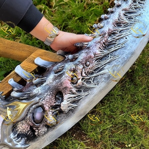 Könnte beinhalten: Ein aufwendiges Schwert mit einer silbernen Klinge und detaillierten Drachenschuppen-Details. Der Knauf hat goldene Akzente und einen großen, lila Edelstein. Das Design ist stark strukturiert und weist aufwendige Schnitzereien auf.
