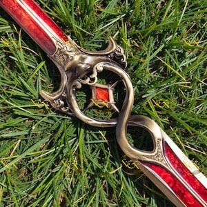 Puede incluir: Una espada decorativa con una hoja roja y dorada, una empuñadura de bronce ornamentada y una gema roja en el centro. La espada está sobre hierba verde, mostrando su intrincado diseño y artesanía.