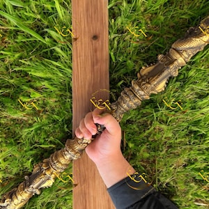 May include: An antique-style staff with a detailed, gold-coloured metal design. The staff is held against a wooden plank, set against a backdrop of green grass. The staff's intricate carvings and overall design suggest a historical or fantasy theme.