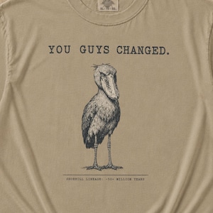 Könnte beinhalten: Ein khakifarbenes Kurzarm-T-Shirt mit einer schwarzen Tuschezeichnung eines Schuhschnabels. Der Text über dem Vogel lautet "YOU GUYS CHANGED." und der Text darunter "SHOEBILL LINEAGE. ~30+ MILLION YEARS." Das T-Shirt hat einen Rundhalsausschnitt.