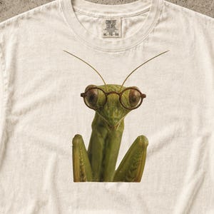 Könnte beinhalten: Ein cremefarbenes Kurzarm-T-Shirt mit einem Grafikdruck einer grünen Gottesanbeterin, die eine runde Brille mit braunem Rand trägt. Das T-Shirt hat einen Rundhalsausschnitt und ein Comfort Colors Markenetikett im inneren Kragenbereich.