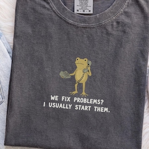 May include: A charcoal gray crew neck t-shirt featuring a graphic of a light green frog holding a telephone handset. Below the frog, the white text reads: WE FIX PROBLEMS? I USUALLY START THEM. The shirt has a Comfort Colors brand label at the neck.