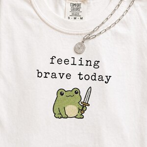 Op de afbeelding: Een crèmekleurig t-shirt met de zwarte tekst 'feeling brave today' boven een afbeelding van een kleine groene kikker die een zilveren zwaard vasthoudt. Een zilverkleurige ketting met een ronde hanger ligt op de halslijn van het shirt.