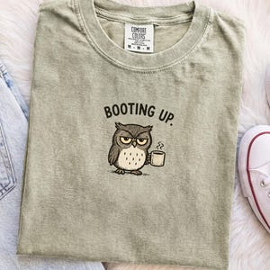 Könnte beinhalten: Ein salbeigrünes T-Shirt mit einer Grafik einer mürrischen Eule, die eine dampfende Kaffeetasse hält, und dem Text BOOTING UP darüber. Das T-Shirt hat einen Rundhalsausschnitt und ein Comfort Colors Markenetikett am inneren Kragen.