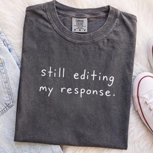 May include: A charcoal gray short-sleeve t-shirt with a crew neckline and the white text "still editing my response." printed on the front. The shirt features a Comfort Colors brand label on the inside collar and is displayed with denim jeans.