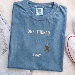 Op de afbeelding: Een blauw t-shirt met ronde hals met de tekst ONE THREAD in lichtgele letters boven een bruine spingrafiek, met het woord AWAY. onder de spin gedrukt. Het shirt heeft een Comfort Colors merklabel aan de binnenkant van de kraag.