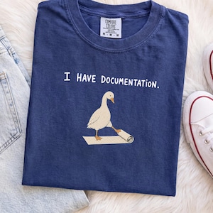 Könnte beinhalten: Ein marineblaues T-Shirt mit Rundhalsausschnitt und einer Grafik einer weißen Gans, die auf einem zusammengerollten Dokument steht. Der Text "I HAVE DOCUMENTATION." ist in Weiß über der Gans gedruckt. Das Shirt hat ein Comfort Colors Etikett.