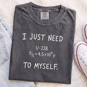 Könnte beinhalten: Ein anthrazitfarbenes Kurzarm-T-Shirt mit weißem Text I JUST NEED U-238 t1/2 = 4.5 x 10^9 y TO MYSELF. Das T-Shirt hat einen Rundhalsausschnitt und ein sichtbares Innenetikett mit der Größe M, platziert neben hellblauen Jeans und weißen Turnschuhen.