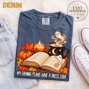 Può includere: Una t-shirt blu con la grafica di un libro aperto, una zucca, una candela accesa e una tazza nera con luna e stelle. Un piccolo fantasma esce dalla tazza e un altro segna una pagina. Il testo recita: My evening plans have a dress code.