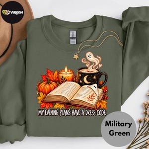 May include: An olive green sweatshirt featuring a graphic of an open book, a lit candle, a mug with a moon design, and a ghost. The text reads, "MY EVENING PLANS HAVE A DRESS CODE." The design includes autumn leaves and berries in orange and red tones.