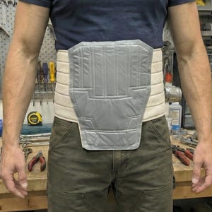 May include: A grey and beige back support with a padded front panel. The support is worn around the waist. The person in the image is wearing a navy blue t-shirt and olive green trousers. The support is designed to provide support.