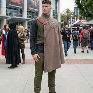 May include: A person in a costume featuring a brown cape, olive green trousers, and black boots. The outfit includes a high-collared top with a quilted design and a red detail. The background shows a convention with posters and other attendees.