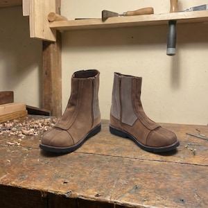 May include: A pair of brown leather ankle boots with a side zipper and a contrasting light brown suede panel. The boots are displayed on a wooden workbench, showcasing their craftsmanship.