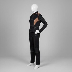 Op de afbeelding: Een zwart tweedelig outfit op een mannequin. De jas heeft bruine accenten en witte stiksels. De broek is zwart met witte stiksels en knielappen. De outfit wordt gepresenteerd op een witte mannequin tegen een grijze achtergrond.