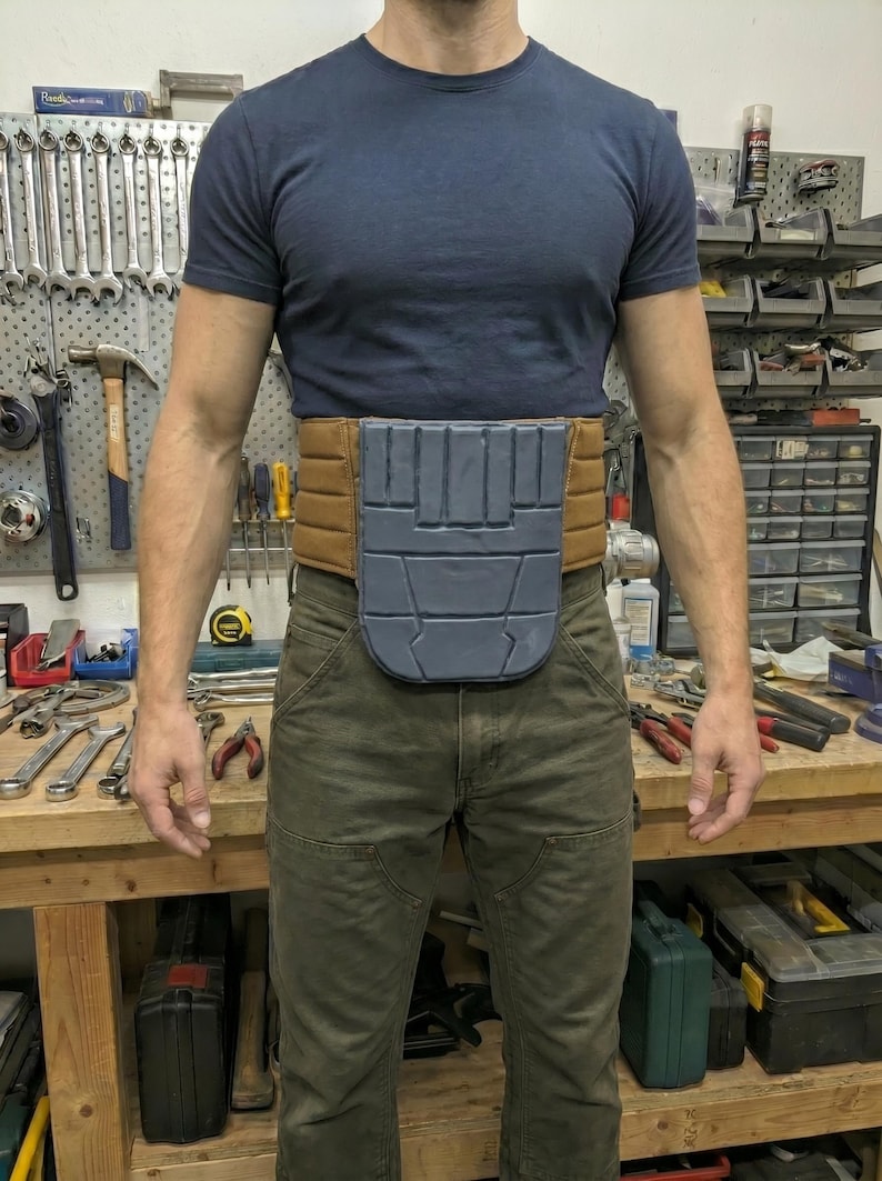May include: A brown and gray utility belt with a gray front panel is worn over green work pants and a navy blue t-shirt. The belt appears to be made of padded material and is designed to hold tools or other items.