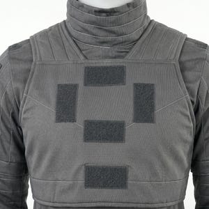 May include: A gray tactical vest with a high collar and multiple hook-and-loop patches. The vest is designed with a padded front and back, and features white stitching details. The overall design suggests a focus on protection and functionality.