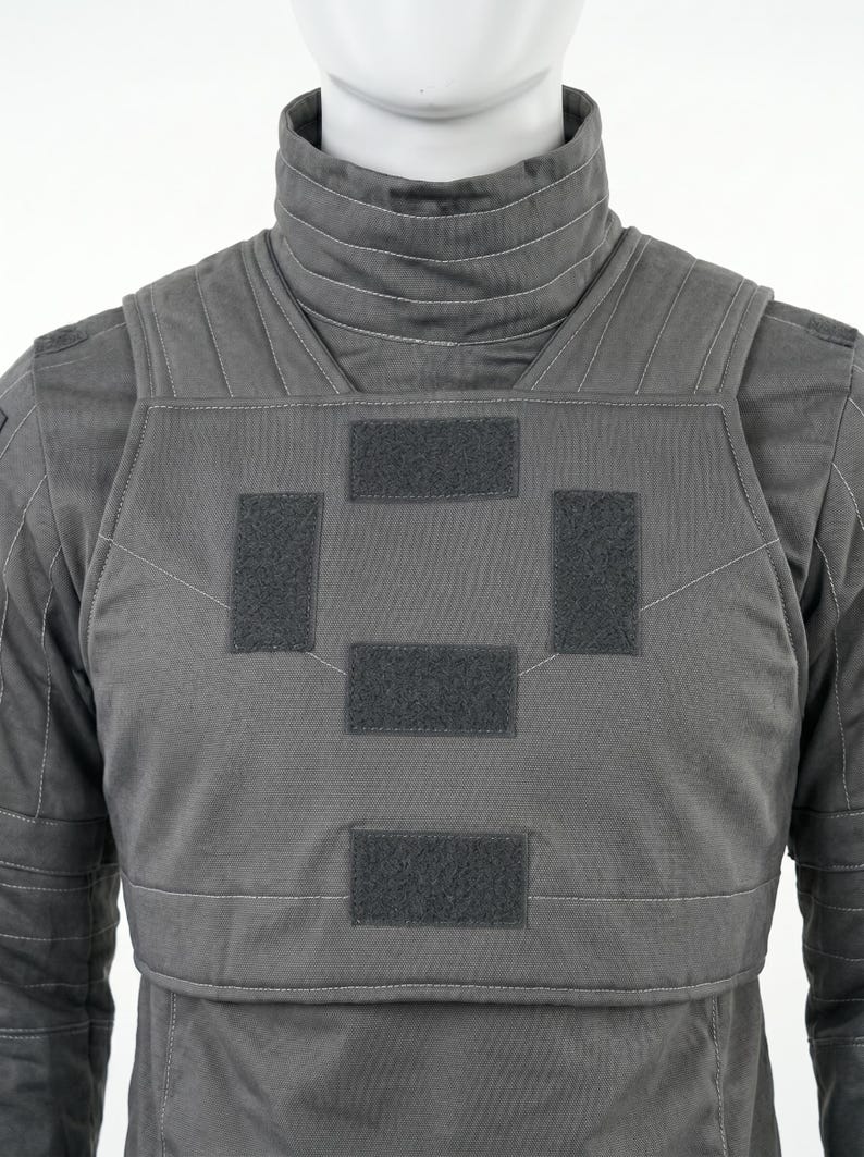 May include: A gray tactical vest with a high collar and multiple hook-and-loop patches. The vest is designed with a padded front and back, and features white stitching details. The overall design suggests a focus on protection and functionality.