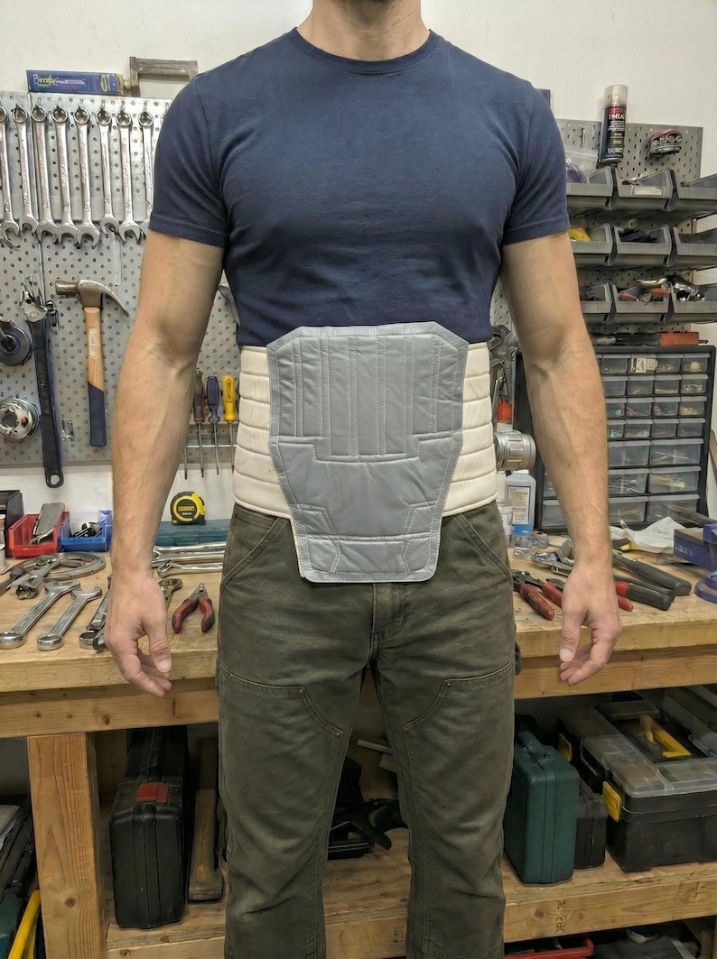 May include: A grey and beige back support with a padded front panel. The support is worn around the waist. The person in the image is wearing a navy blue t-shirt and olive green trousers. The support is designed to provide support.