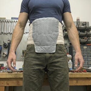 May include: A grey and beige back brace with a quilted design. The brace is worn around the waist and features a padded section for back support. The person is wearing a blue t-shirt and green trousers.