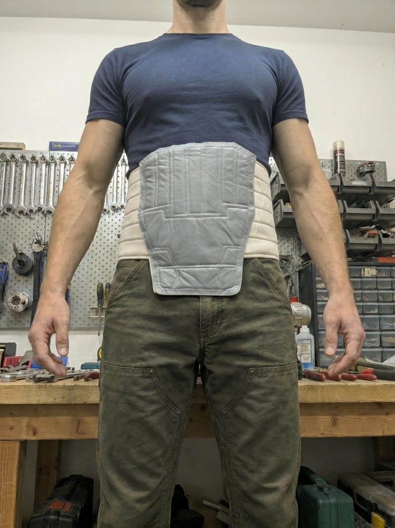 May include: A grey and beige back brace with a quilted design. The brace is worn around the waist and features a padded section for back support. The person is wearing a blue t-shirt and green trousers.