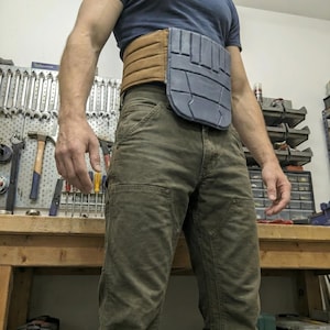 May include: A brown and gray utility belt with a segmented design. The belt is worn over a blue t-shirt and olive green pants. The belt appears to be made of leather and a hard plastic material.