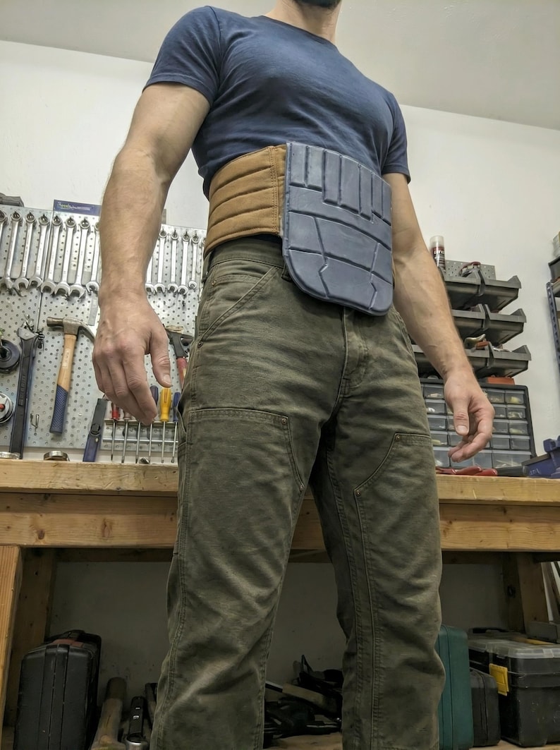 May include: A brown and gray utility belt with a segmented design. The belt is worn over a blue t-shirt and olive green pants. The belt appears to be made of leather and a hard plastic material.