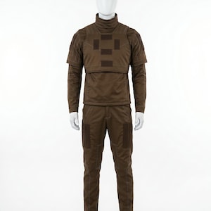 Pode incluir: Um uniforme t&aacute;tico castanho exibido em um manequim. O conjunto inclui uma parte superior de manga comprida com gola alta e peitoral, e cal&ccedil;as a condizer. Apresenta remendos e costuras refor&ccedil;adas.