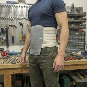May include: A grey and beige padded support belt worn around the waist. The belt has a rectangular grey section on the front and is worn over a dark blue t-shirt and green trousers. Tools are visible in the background.