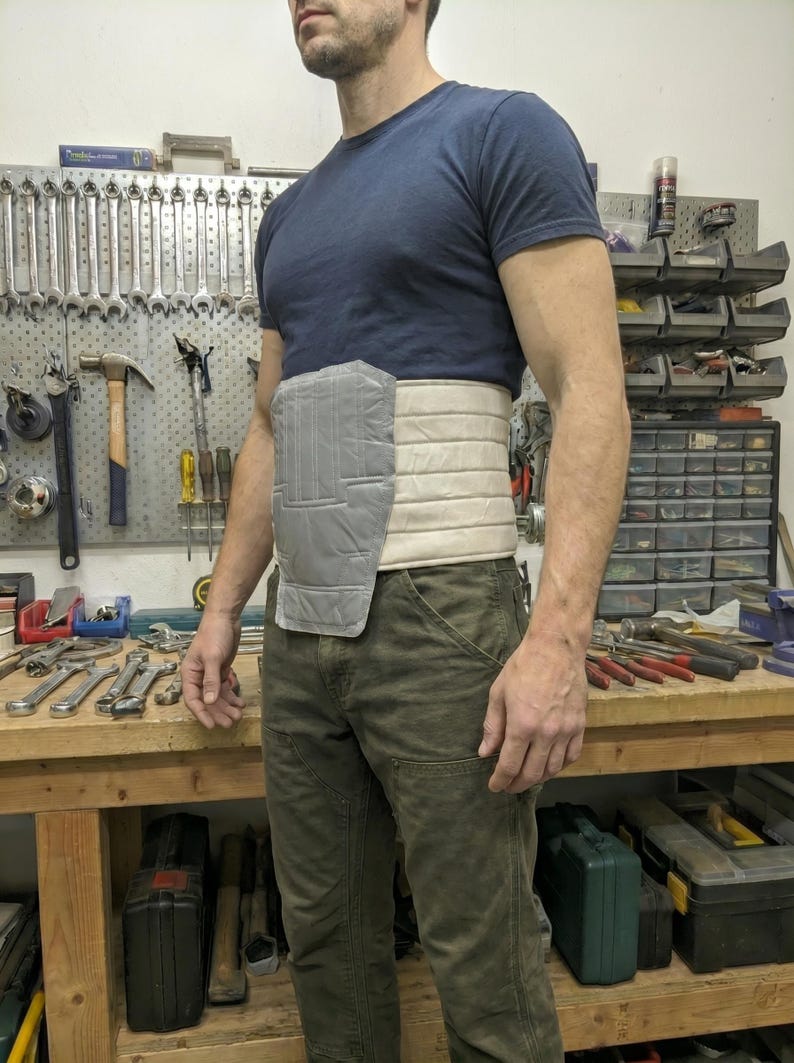 May include: A grey and beige padded support belt worn around the waist. The belt has a rectangular grey section on the front and is worn over a dark blue t-shirt and green trousers. Tools are visible in the background.