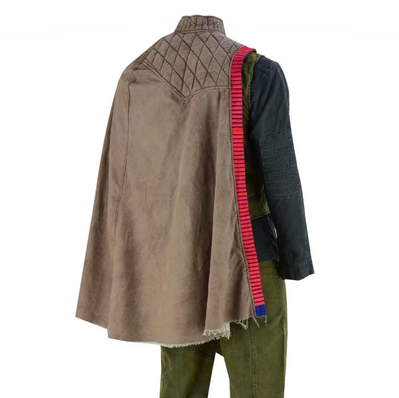 May include: A brown cape with a quilted shoulder detail and a red and blue strap. The cape is paired with a dark green and black jacket and green trousers. The outfit has a distressed, worn appearance.