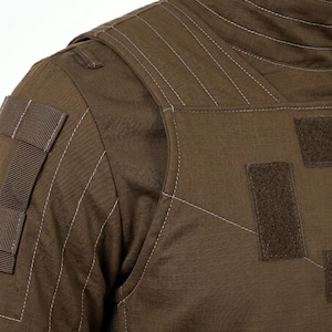 Pode incluir: Close-up de um colete t&aacute;tico castanho com detalhes de costura branca. O colete apresenta v&aacute;rios patches e correias de velcro, mostrando um design dur&aacute;vel e funcional. O tecido tem uma apar&ecirc;ncia texturizada.