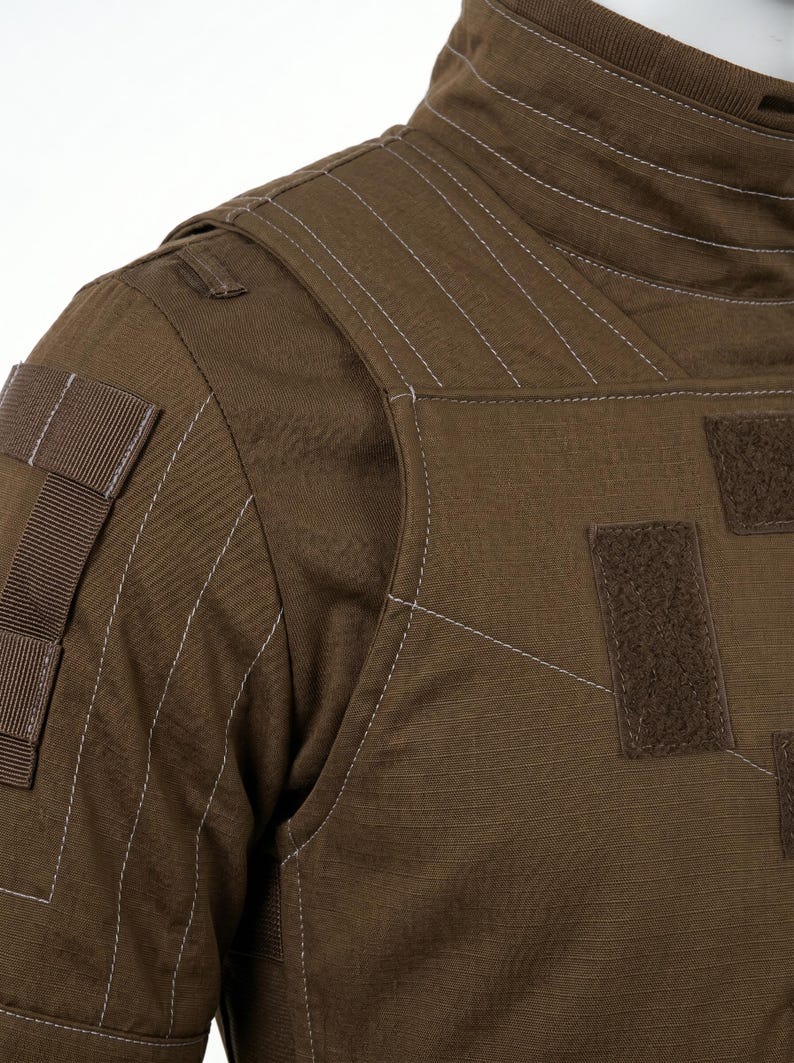 Pode incluir: Close-up de um colete t&aacute;tico castanho com detalhes de costura branca. O colete apresenta v&aacute;rios patches e correias de velcro, mostrando um design dur&aacute;vel e funcional. O tecido tem uma apar&ecirc;ncia texturizada.