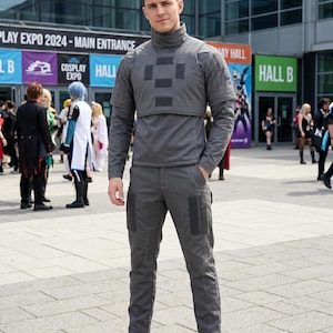 May include: A person wearing a gray cosplay outfit, including a high-neck top, pants, and boots. The outfit has dark gray patches on the chest and legs. The background includes signs for a cosplay expo.