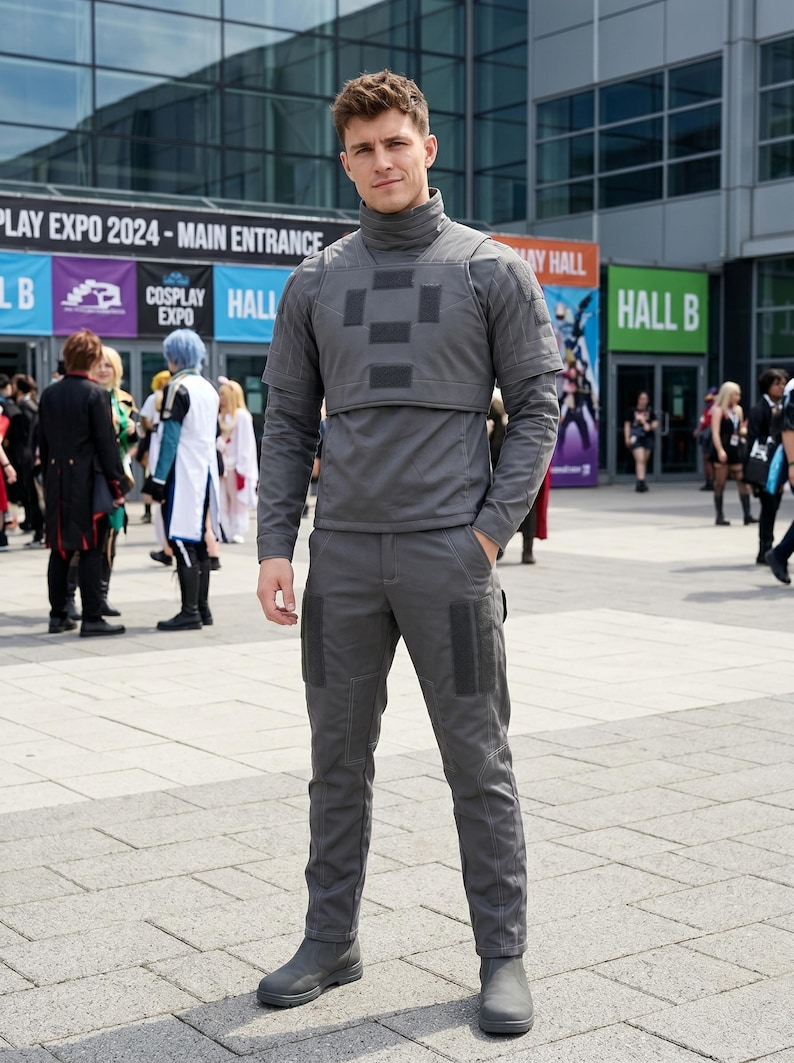 May include: A person wearing a gray cosplay outfit, including a high-neck top, pants, and boots. The outfit has dark gray patches on the chest and legs. The background includes signs for a cosplay expo.