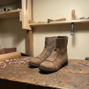 May include: Two brown leather ankle boots with zippers, displayed on a wooden workbench. The boots have a rugged, worn appearance, with a dark sole and a cap toe design. The background shows woodworking tools and a wooden shelf.