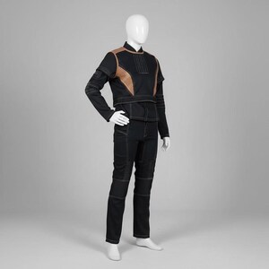 Op de afbeelding: Zwart jumpsuit met bruine accenten en witte stiksels. De outfit bestaat uit een jas met lange mouwen en een bijpassende broek. De jas heeft bruine schouderpanelen en een hoge kraag. De broek heeft versterkte kniepanelen.