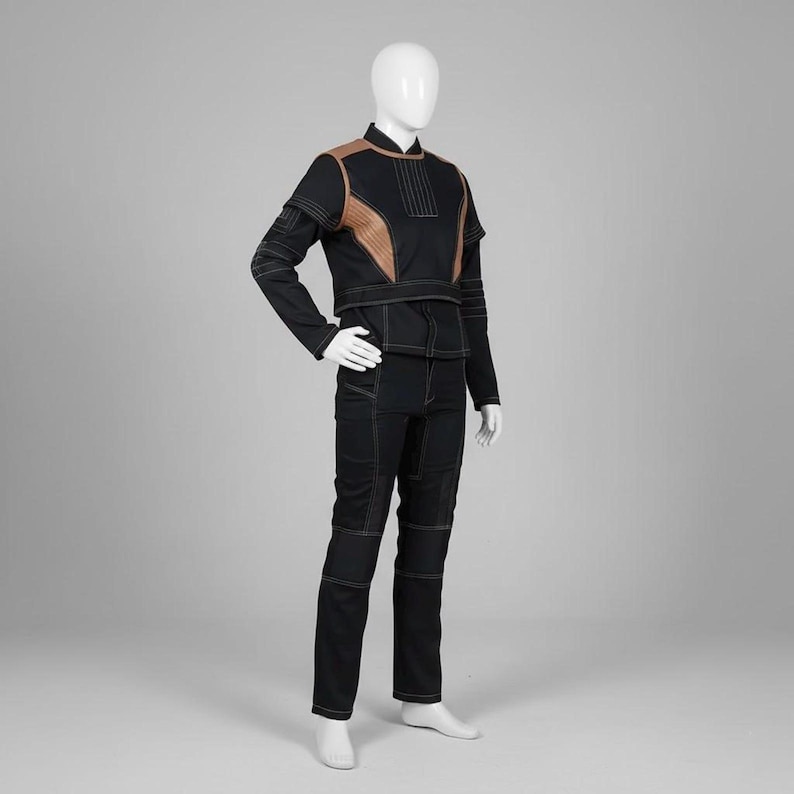 Op de afbeelding: Zwart jumpsuit met bruine accenten en witte stiksels. De outfit bestaat uit een jas met lange mouwen en een bijpassende broek. De jas heeft bruine schouderpanelen en een hoge kraag. De broek heeft versterkte kniepanelen.