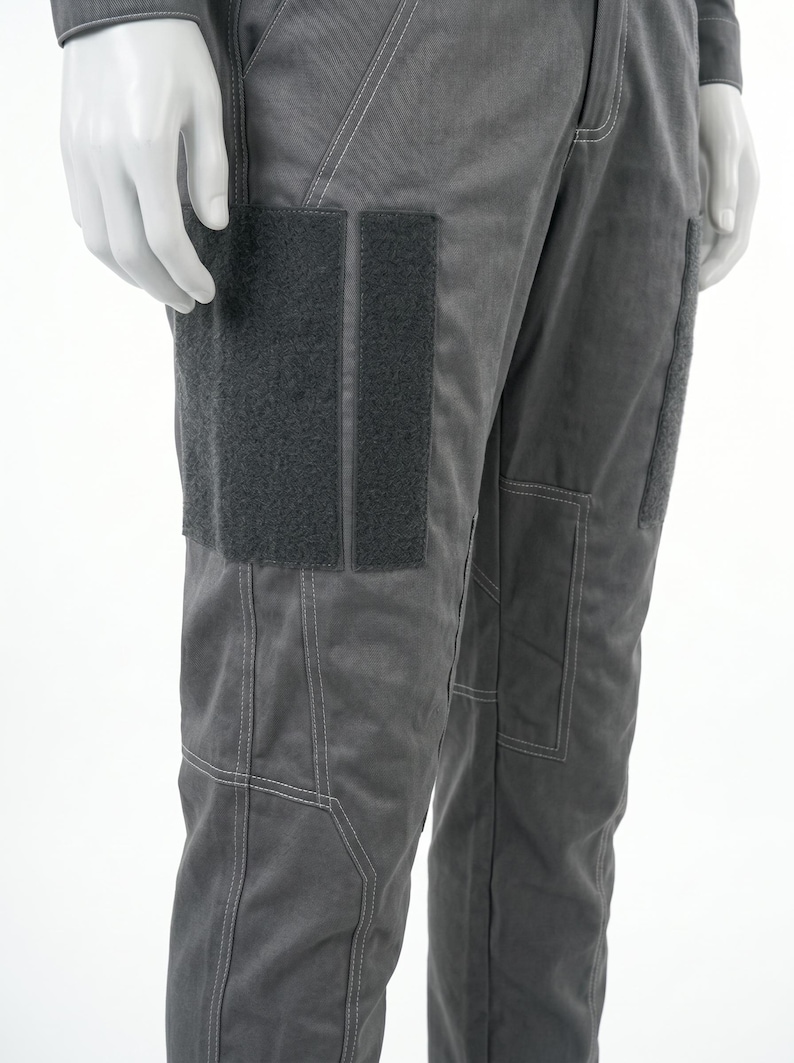May include: Close-up of gray pants with visible stitching and two dark gray rectangular patches on the left thigh. The pants have a utilitarian design, with a pocket on the right thigh. The fabric appears to be a durable, possibly workwear material.