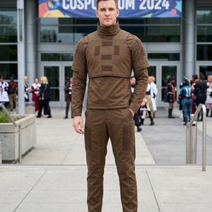 Pode incluir: Uma pessoa vestindo uma roupa de cosplay marrom com gola alta e patches quadrados. A roupa inclui uma blusa de manga comprida, cal&ccedil;as e botas. O fundo apresenta um pr&eacute;dio com uma faixa que diz "COSPORUM 2024".