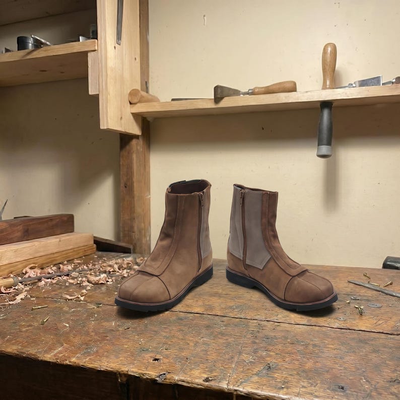 May include: A pair of brown leather ankle boots with a side zipper and a contrasting light brown suede panel. The boots are displayed on a wooden workbench, showcasing their craftsmanship.