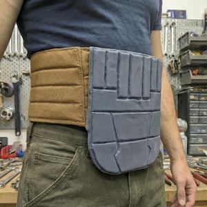 May include: A brown and gray utility belt worn around the waist. The belt has a padded brown section and a gray, rectangular section with a geometric design. The person is wearing a blue shirt and green pants.