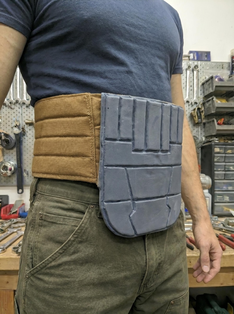 May include: A brown and gray utility belt worn around the waist. The belt has a padded brown section and a gray, rectangular section with a geometric design. The person is wearing a blue shirt and green pants.