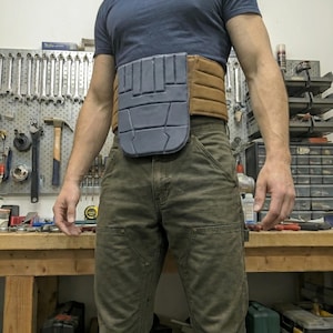 May include: A utility belt with a gray front panel and a brown belt section. The belt is worn over green work pants and a blue t-shirt. The background shows a workbench with tools.