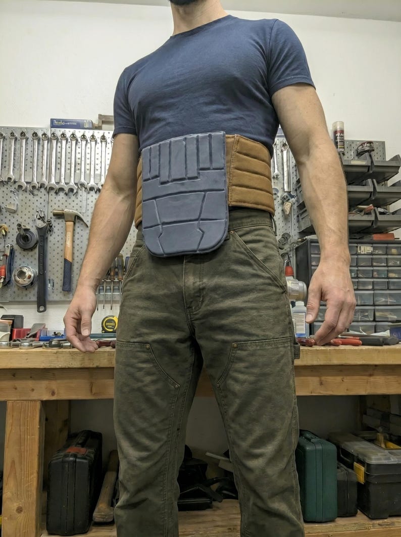 May include: A utility belt with a gray front panel and a brown belt section. The belt is worn over green work pants and a blue t-shirt. The background shows a workbench with tools.