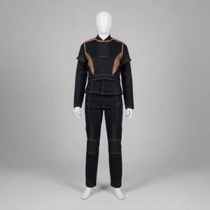 Op de afbeelding: Een zwart tweedelig outfit op een mannequin. De top heeft een hoge kraag en koperkleurige accenten op de schouders en borst. De broek is zwart met witte stiksels. De outfit is een futuristisch ontwerp.